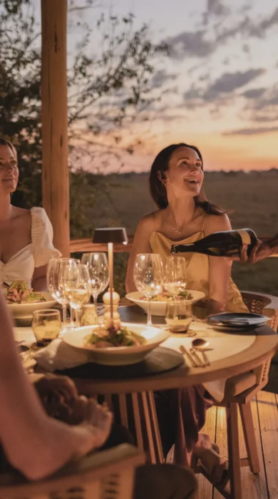 Three women sit around dinner table as the waiter pours wine, Mombo, Moremi Game Reserve, Botswana - Yellow Zebra Safari