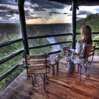 A guest sits on a wooden decking and looks out on the river, Imvelo Safari Lodge, Hwange National Park, Zimbabwe - Yellow Zebra Safari