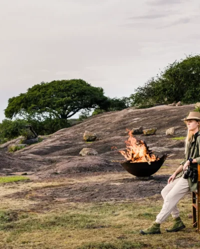 Aside a firepit, woman lounges on a camp chair in a rocky area - Yellow Zebra Safaris